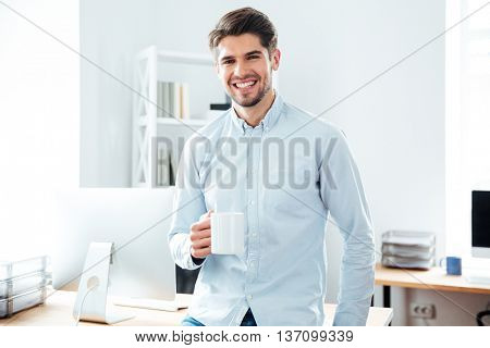 Smiling young businessman standing and drinkig coffee in office