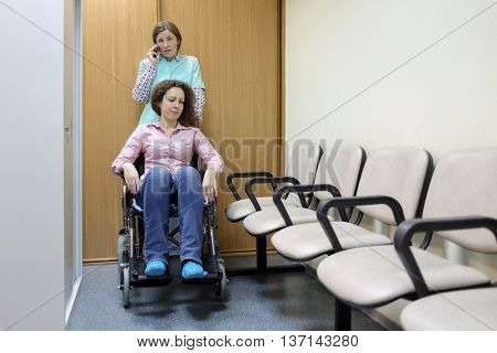 Nurse talking on phone in hospital corridor, standing next to patient in wheelchair, holding handles