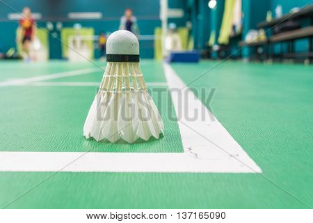 White badminton shuttlecock on a green floor with blurred players in badminton court.