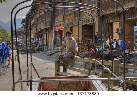 Zhaoxing Dong Village Guizhou Province China - April 8 2010: Asian man sitting on a bench in the middle of the rural street near the wooden houses with shops and restaurants a spring sunny day.