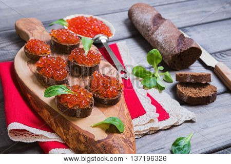 Small canape sandwiches with fish red caviar leaves of green salad rye bagget red linen napkin with lace on cutting board olive wood gray background wooden surfaces