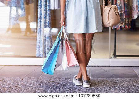 Closeup of a young woman's legs standing in front of a clothing store holding many shoppping bags and her handbag, standing in front of a store window