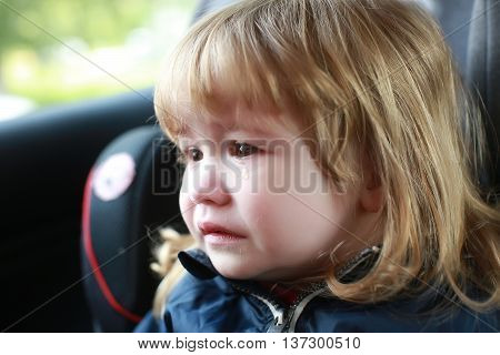 small boy child with facial emotions on unhappy crying face and long blonde hair sitting in car with tear on cheek closeup
