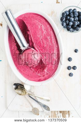 Homemade bluberry ice cream and scooper in mold served with fresh berries, silver spoons on white rusric wooden background, top view
