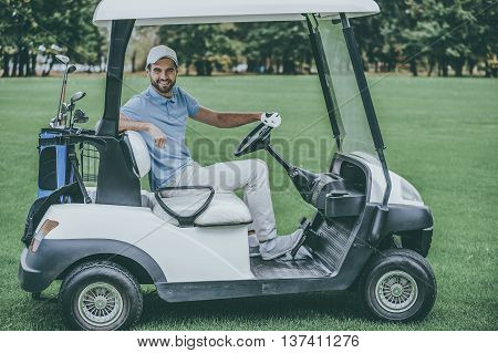 Driving golf cart. Side view of handsome young man driving a golf cart and looking at camera