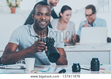 Confident photographer. Handsome young African man holding camera and smiling while three people working on background