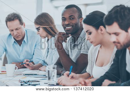 Happy to be a part of grat team. Group of business people sitting in a row at the table while handsome African man looking at camera and smiling