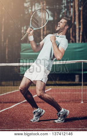 Celebrating victory. Full length of happy young man in polo shirt holding tennis racket and gesturing while standing on tennis court