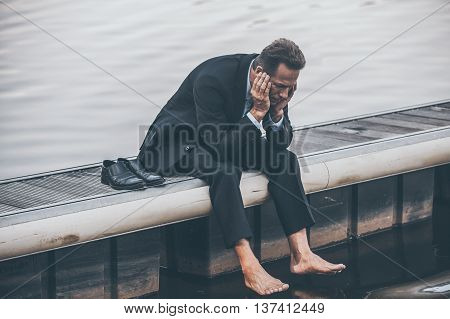 Depressed and hopeless. Depressed mature businessman holding head in hands while sitting barefoot at the quayside