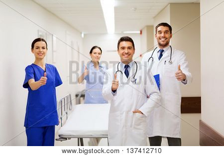 clinic, profession, people, health care and medicine concept - group of happy medics or doctors with gurney showing thumbs up at hospital corridor