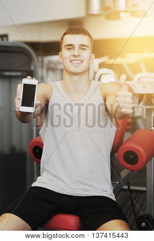 sport, bodybuilding, lifestyle, technology and people concept - smiling young man showing smartphone and thumbs up gesture in gym