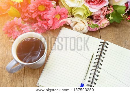 Open a blank white notebook, pen and cup of coffee on the desk with flower background under morning sunlight