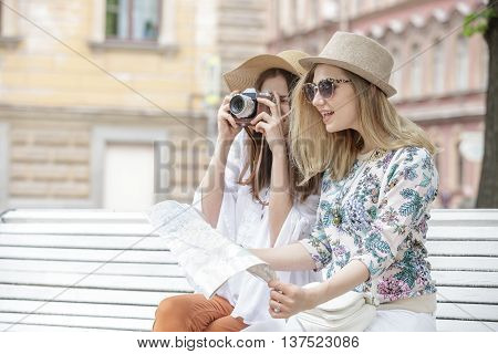 Beautiful girls tourists are looking for an address on the map sitting on the bench. They are wearing hats and smiling. Outdoors. Summer day.