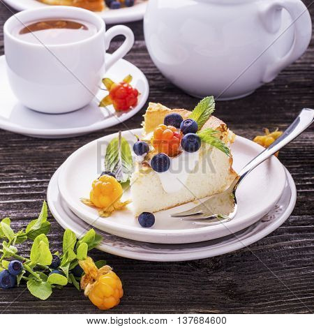 Homemade breakfast. Fresh cheesecake with Nordic berries cloudberries and blueberries in plain wooden background served with cloudberry tea in a white ceramic bowl. Decorated branches forest blueberries