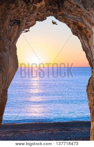 Deep high Rock Cave on Seafront and extreme Climbers Sea and Rising Sun on Background