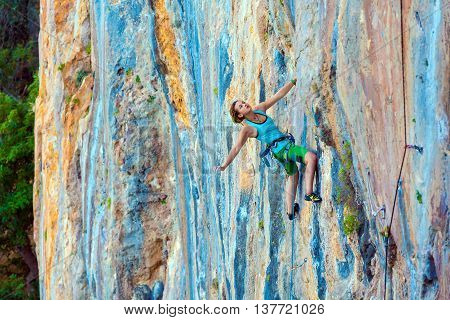 Young Female Climber ascending vertical rocky wall sporty Clothing Blue Shirt Green Pants using Rope and other Safety Gear