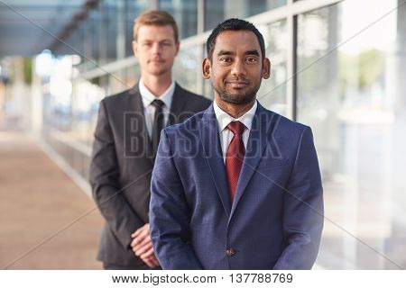 Portrait of a confident and successful businessman standing in front of the glass windows of a modern office building with a colleague in the background