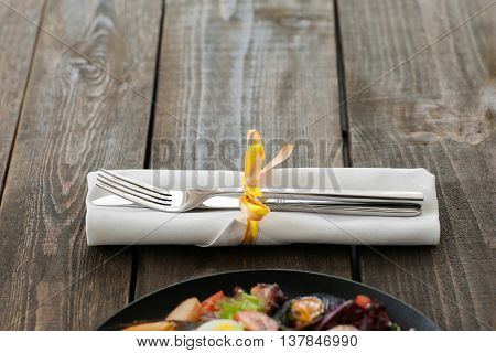 Cutlery on brown wooden background with seafood, void. Horizontal position of white napkin with knife and fork, seafood meal in downside of image, copyspace on wood. Focus on restaurant cutlery