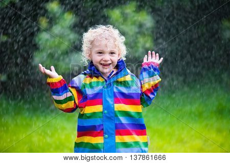 Little blond curly boy in a waterproof jacket in rainbow color playing in the rain. Kids having fun outdoors in autumn shower.