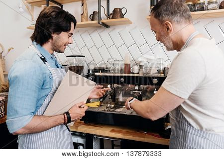Making coffee together. Happy older male barista showing his younger positive and merry colleague how to use coffee machine