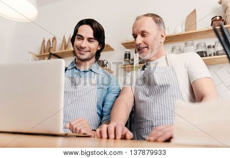 Technologies at work. Cheerful and positive two male cafe workers using laptop at work
