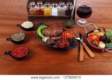 fresh roast beef meat rib eye steak with tomatoes,potatoes, hot chili pepper gern and pink peppercorn saucer, cutlery, set of spice, and red wine glass on light walnut table
