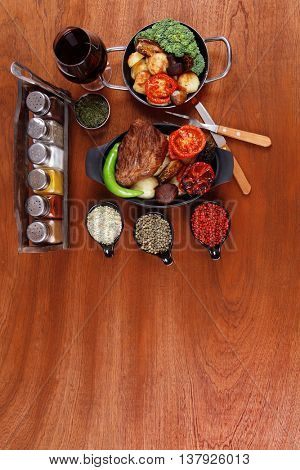 fresh roast beef meat rib eye steak with tomatoes,potatoes, hot chili pepper gern and pink peppercorn saucer, cutlery, set of spice, and red wine glass on light walnut table