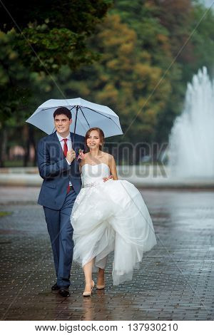 Happy wedding couple in a rainy wedding day outdoors in park with white umbrella in rainy weather. Bride and groom are embracing and walking in park.