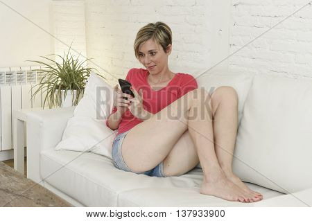 young beautiful Caucasian woman with shorts and modern hair style happy on couch using internet texting on mobile phone relaxed and cheerful at home living room in communication concept