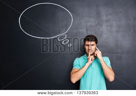 Handsome young man talking on cell phone and showing silence gesture with thinking bubble on chalkboard background
