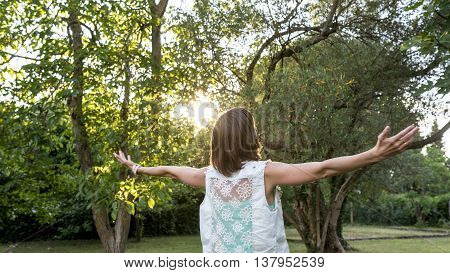 Young woman celebrating the morning sun and spring season as she stands outdoors in the garden at sunrise with outspread arms rear view.