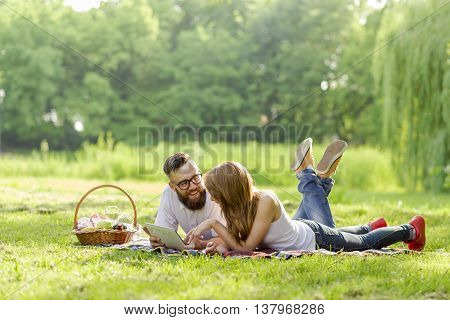 Couple lying on a picnic blanket in a park with a picnic basket filled with fruit food and wine placed next to them surfing the net on a tablet computer