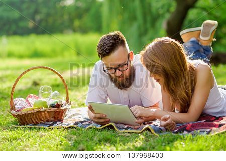 Couple lying on a picnic blanket in a park with a picnic basket filled with fruit food and wine placed next to them surfing the net on a tablet computer