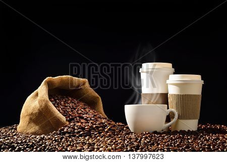 Cups of coffee with smoke and coffee beans in burlap sack on black background