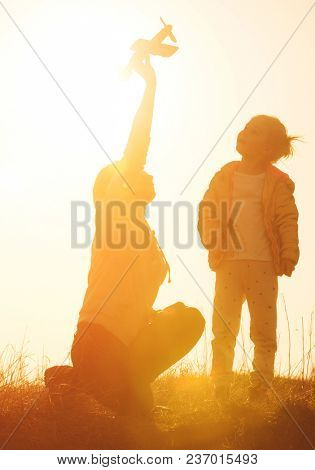 Silhouette of mother and daughter playing in the field with small airplane toy against vivid orange sky. Happy mother and daughter at sunset time