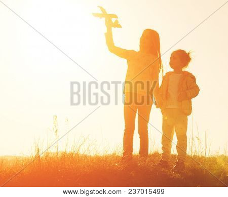 Two little sisters holding hands and playing with small airplane toy outdoors. Silhouette of two sisters walking on the meadow at sunset