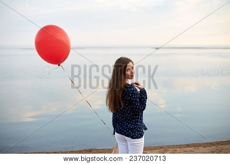 Portrait Of Young Woman With Red Air Balloon And Present Bag Near The Calm Sea Or Lake. Clouds Are R