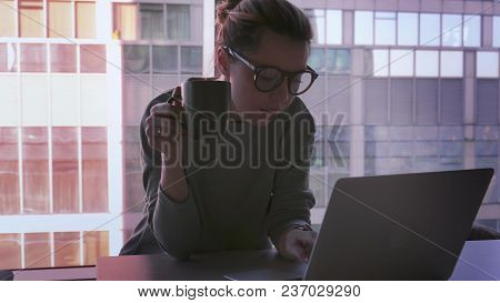 Business Woman Is Sitting In Office With Laptop, Drinking Coffee, Smiling And Laughing. Skyscrapers 