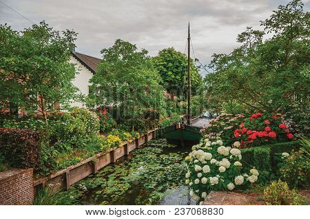 Charming Small Canal With Boat Next To A Rustic House And Lush Flowery Garden In Cloudy Day At Drimm