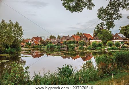 Charming Wide Canal Next To Rustic Houses With Green Bushes And Trees Reflected On The Water In Clou