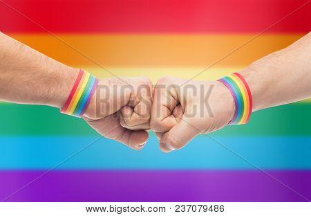 lgbt, same-sex love and homosexual relationships concept - close up of male couple hands with gay pride awareness wristbands making fist bump gesture over rainbow background