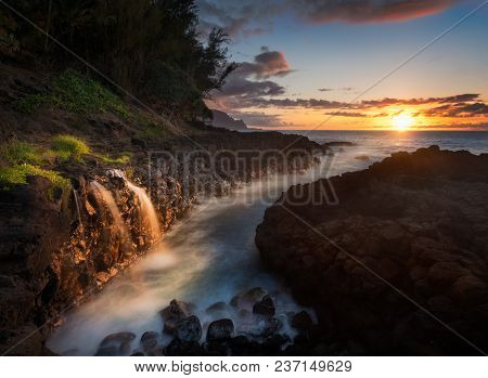 Setting Sun At Sunset Illuminates A Small Waterfall Falling Into The Ocean With The Na Pali Coastlin
