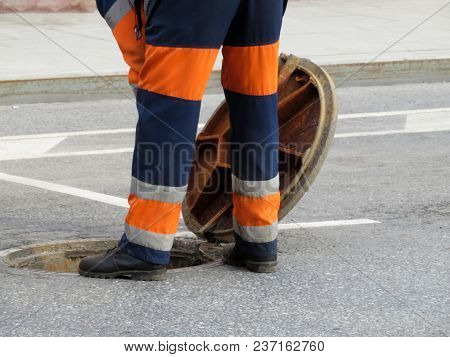 The Worker In Uniform Stand Over The Open Sewer Hatch. Repair Of Sewage Or Underground Utilities