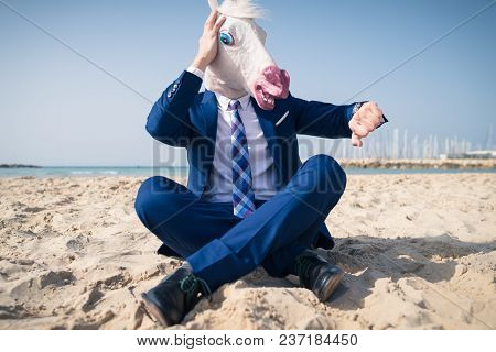 Stylish Manager In Comical Mask And Elegant Suit Sits On Beach And Looks On The Watch. Scared Man Lo