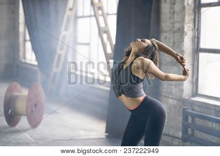 Charming Girl With Closed Eyes In A Dark Sportswear Engaged In Yoga On The Windows Background In The