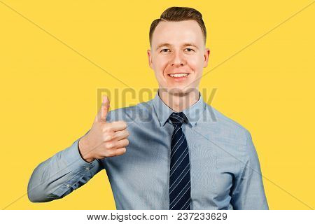 Young Businessman Dressed In Blue Shirt And Tie Shows Thumb Up, Isolated On Yellow Background