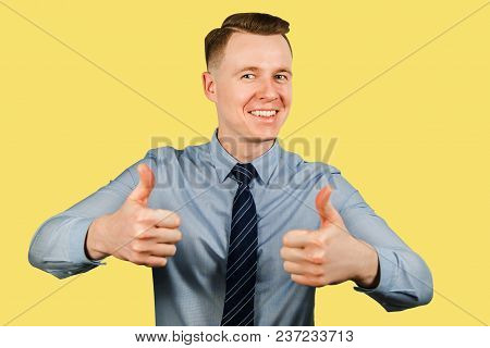 Young Businessman Dressed In Blue Shirt And Tie Shows Thumbs Up And Smiles, Isolated On Yellow Backg