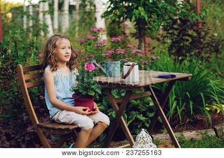 Happy Kid Girl Playing In Summer Garden, Holding Heranium Flower In Pot. Teaching Kids To Love Natur
