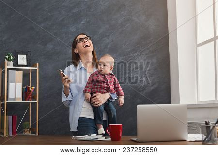 Laughing Mother Working At Office With Her Baby. Young Woman Talking On Phone And Using Laptop While