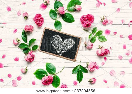 Flowers Composition. Frame Made Of Fresh Rose Flowers With Heart On Chalkboard On White Wooden Backg
